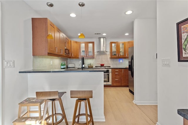 kitchen with glass insert cabinets, pendant lighting, brown cabinetry, and wall chimney exhaust hood