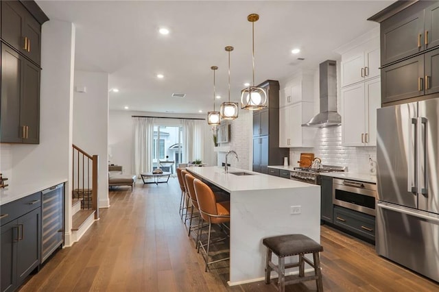 kitchen featuring white cabinetry, a kitchen island with sink, stainless steel appliances, beverage cooler, and wall chimney exhaust hood