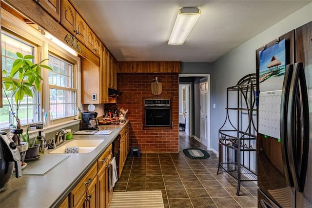 kitchen with dark tile patterned floors, a sink, brown cabinetry, black appliances, and under cabinet range hood