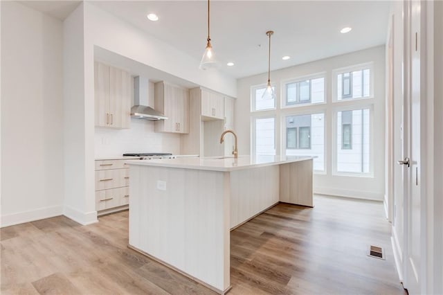 kitchen featuring decorative light fixtures, sink, wall chimney range hood, a center island with sink, and light wood-type flooring