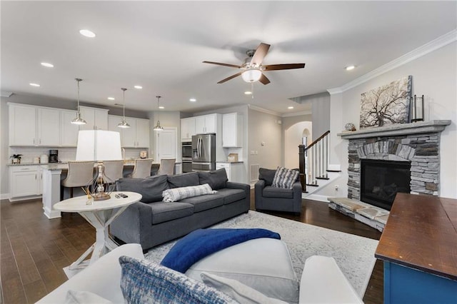 living room with arched walkways, ceiling fan, a stone fireplace, dark wood-style flooring, and crown molding