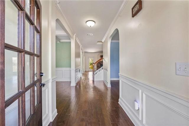 corridor with crown molding, dark wood finished floors, a decorative wall, stairway, and wainscoting