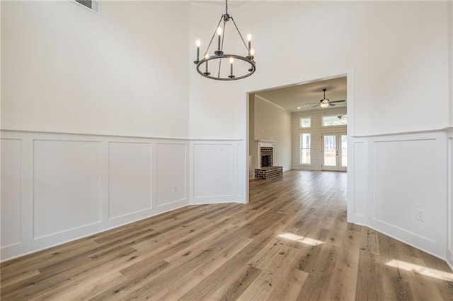 unfurnished dining area with light wood finished floors, visible vents, a brick fireplace, ceiling fan with notable chandelier, and a decorative wall