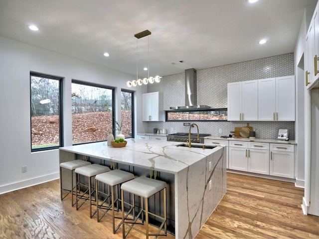 kitchen with light hardwood / wood-style flooring, wall chimney exhaust hood, light stone countertops, an island with sink, and white cabinetry