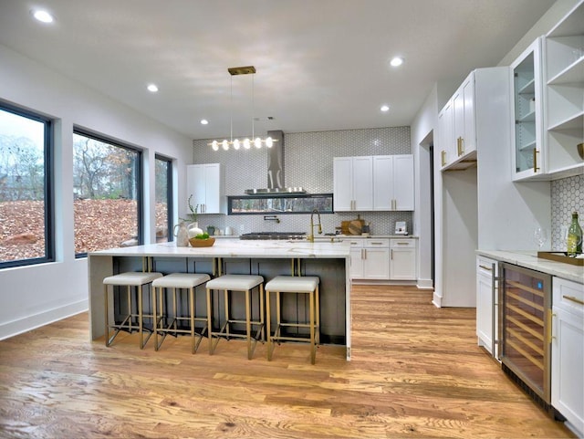 kitchen featuring light wood-type flooring, beverage cooler, a center island with sink, white cabinets, and hanging light fixtures