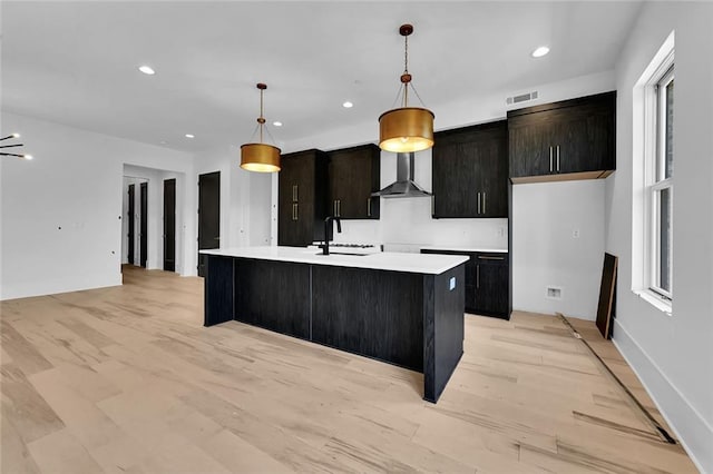 kitchen with wall chimney exhaust hood, an island with sink, decorative light fixtures, and light hardwood / wood-style floors