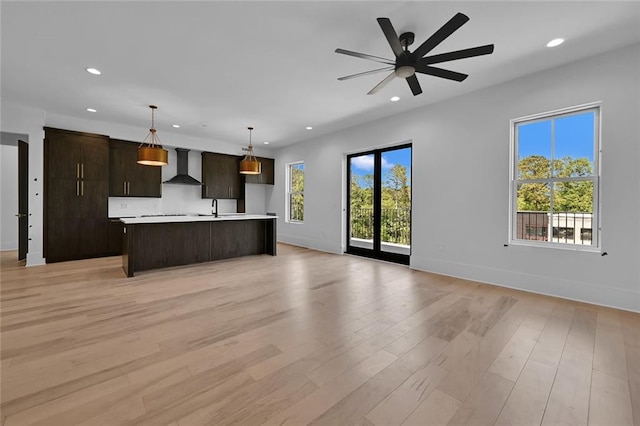 kitchen featuring light wood-type flooring, an island with sink, ceiling fan, and wall chimney range hood