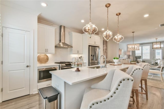 kitchen with an inviting chandelier, a sink, wall chimney range hood, light wood-style floors, and appliances with stainless steel finishes