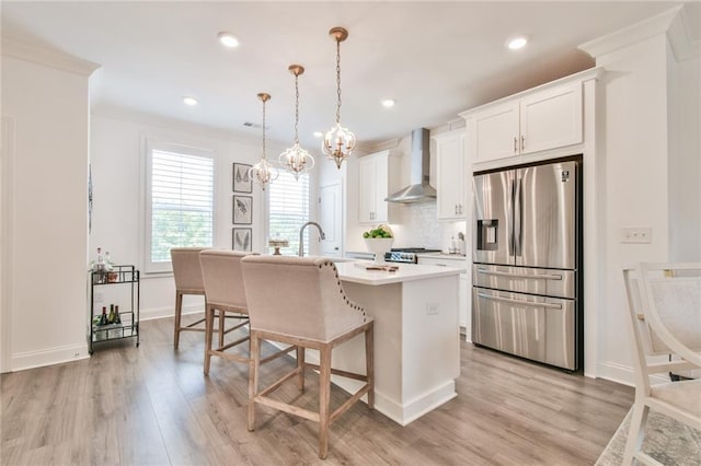 kitchen featuring light countertops, stainless steel fridge, a kitchen island with sink, light wood-type flooring, and wall chimney range hood