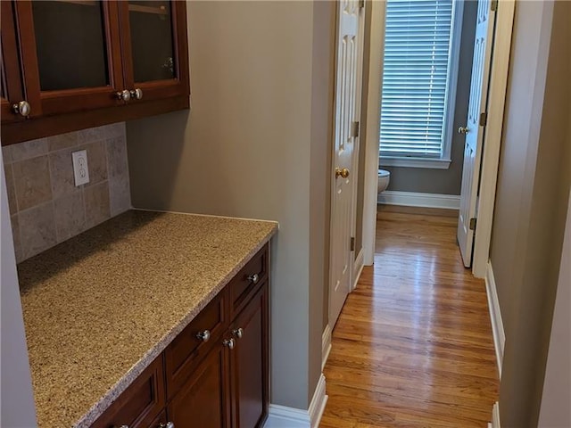 interior space featuring light stone counters, light wood finished floors, backsplash, glass insert cabinets, and baseboards