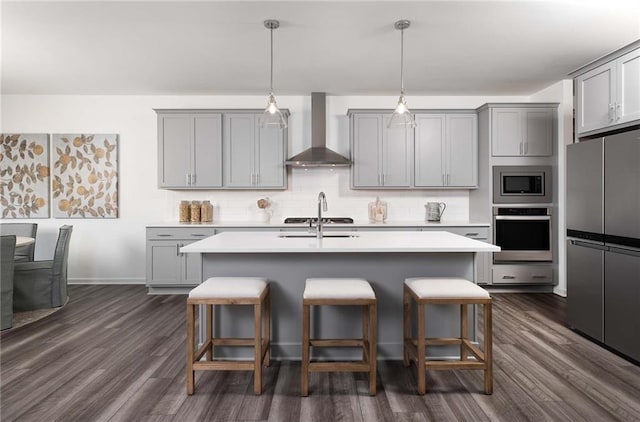 kitchen featuring tasteful backsplash, stainless steel appliances, wall chimney range hood, decorative light fixtures, and an island with sink