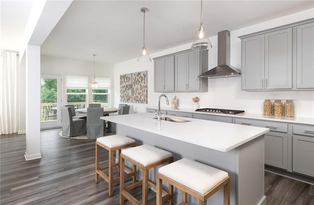 kitchen with sink, hanging light fixtures, wall chimney range hood, stainless steel gas cooktop, and a center island with sink