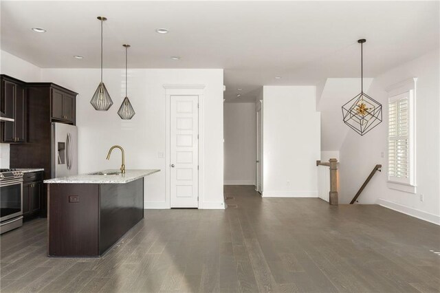 kitchen with wall chimney exhaust hood, hanging light fixtures, dark brown cabinetry, a kitchen island with sink, and appliances with stainless steel finishes