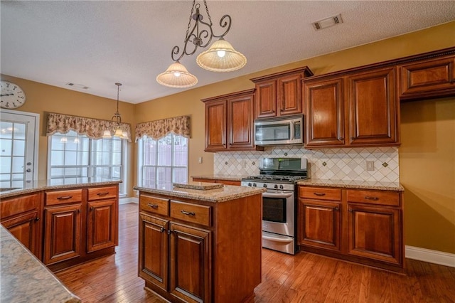 kitchen with appliances with stainless steel finishes, decorative backsplash, hanging light fixtures, light hardwood / wood-style floors, and light stone countertops