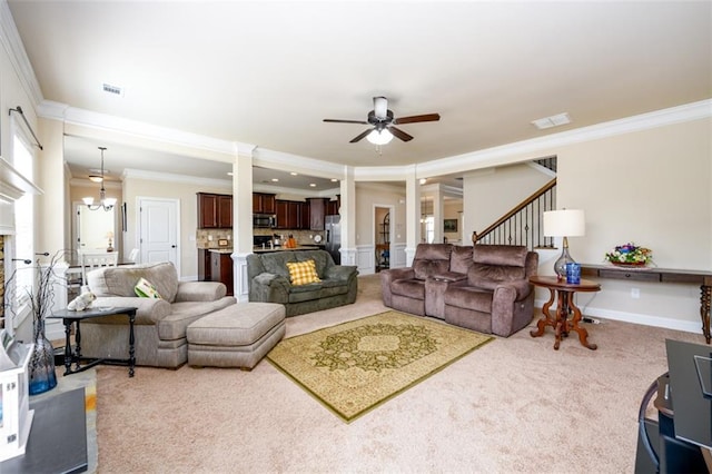 carpeted living area with visible vents, decorative columns, stairs, crown molding, and ceiling fan with notable chandelier