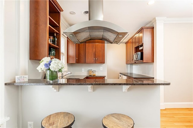 kitchen with open shelves, stainless steel microwave, island range hood, dark stone counters, and a kitchen breakfast bar