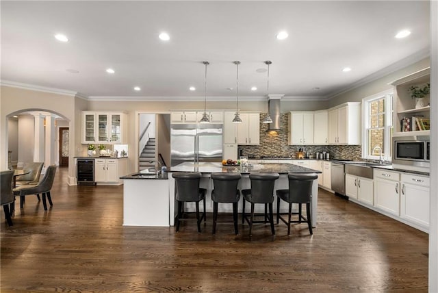 kitchen with built in appliances, arched walkways, beverage cooler, wall chimney range hood, and an island with sink