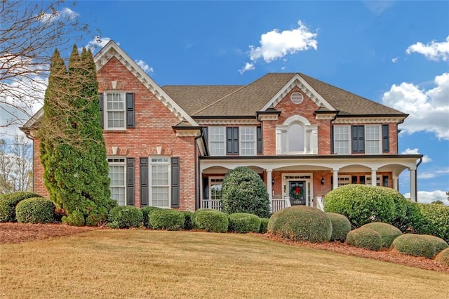 view of front of property featuring covered porch, a front lawn, roof with shingles, and brick siding