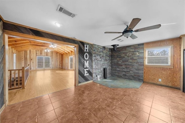 unfurnished living room with a wood stove, wooden walls, a ceiling fan, and visible vents