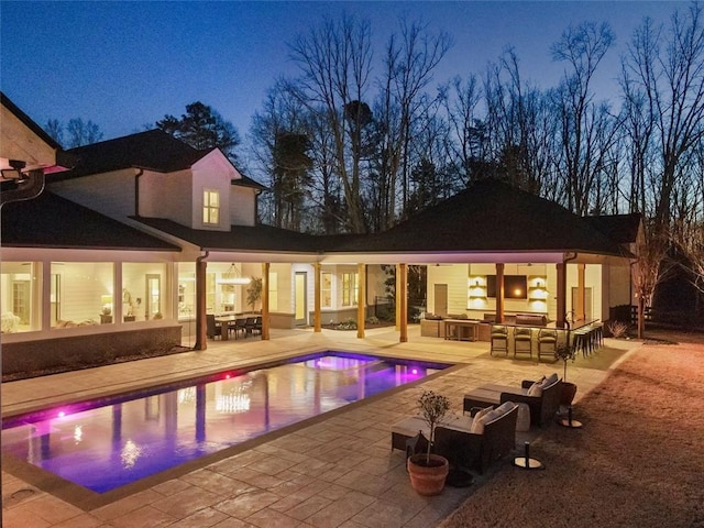 pool at dusk featuring a bar, a patio area, and exterior kitchen