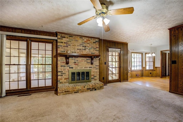 unfurnished living room with a textured ceiling, light carpet, wood walls, a brick fireplace, and ceiling fan with notable chandelier