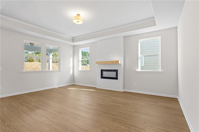 unfurnished living room with a tray ceiling, crown molding, baseboards, a glass covered fireplace, and light wood-style floors