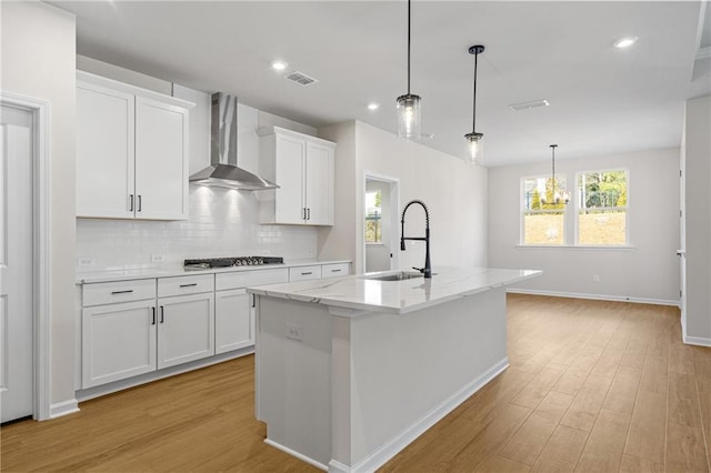 kitchen with a sink, wall chimney exhaust hood, an island with sink, light wood finished floors, and gas stovetop