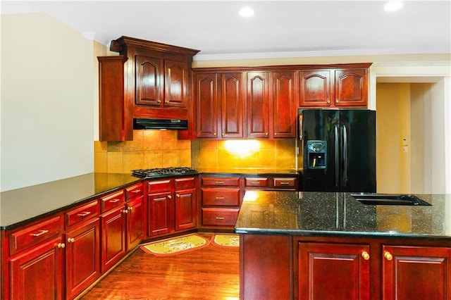 kitchen with gas stovetop, under cabinet range hood, reddish brown cabinets, backsplash, and black refrigerator with ice dispenser