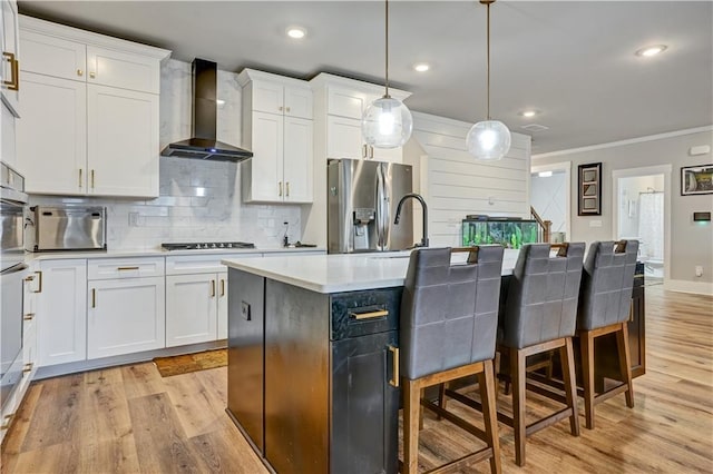 kitchen with white cabinetry, an island with sink, hanging light fixtures, stainless steel fridge, and wall chimney range hood