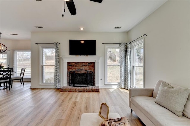 living room with visible vents, light wood-style floors, a brick fireplace, and ceiling fan with notable chandelier