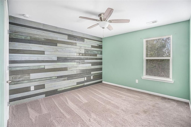 carpeted empty room with an accent wall, wood walls, baseboards, visible vents, and ceiling fan