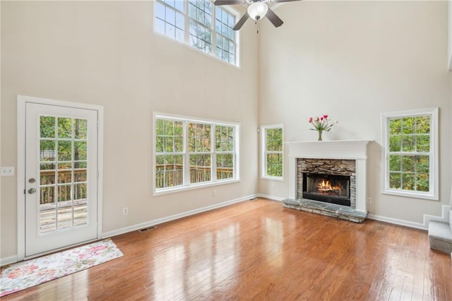 unfurnished living room with ceiling fan, a stone fireplace, a high ceiling, and hardwood / wood-style floors