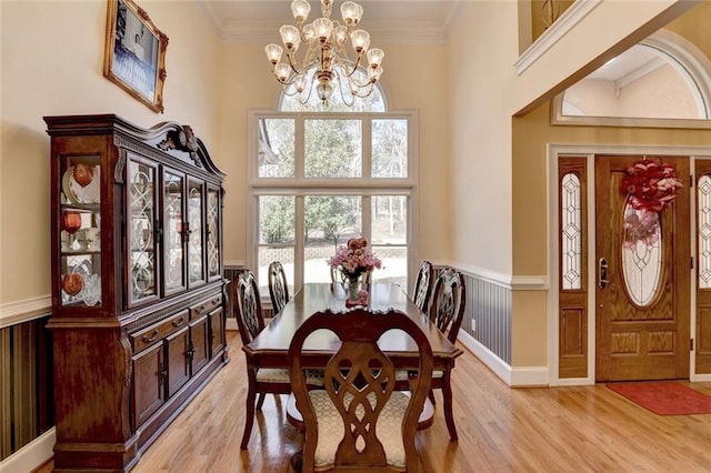 dining area with a wainscoted wall, light wood-style flooring, ornamental molding, an inviting chandelier, and a high ceiling