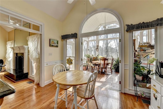 dining space featuring ceiling fan, light wood-style flooring, high vaulted ceiling, and a fireplace with raised hearth