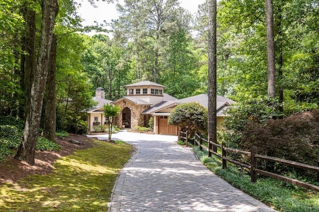 view of front facade with fence, stone siding, decorative driveway, a chimney, and a view of trees