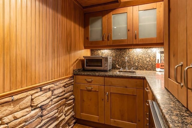 kitchen with a toaster, dark stone counters, glass insert cabinets, brown cabinets, and a sink