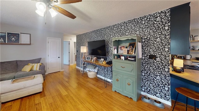 living area with wallpapered walls, a ceiling fan, light wood finished floors, and a textured ceiling