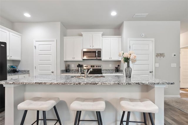 kitchen with stainless steel appliances, wood finished floors, a kitchen island with sink, white cabinets, and a breakfast bar area