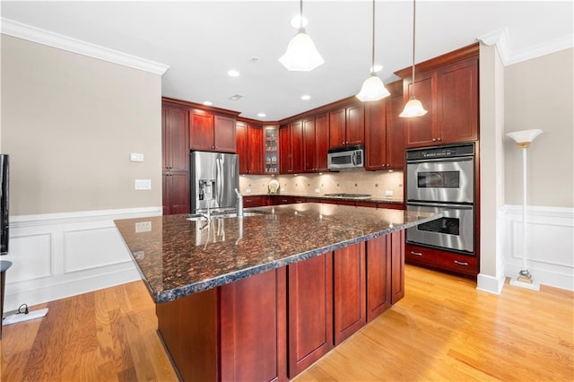 kitchen featuring appliances with stainless steel finishes, backsplash, decorative light fixtures, a center island with sink, and light hardwood / wood-style floors