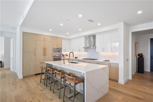 kitchen with light brown cabinets, wall chimney exhaust hood, light hardwood / wood-style floors, and a center island with sink