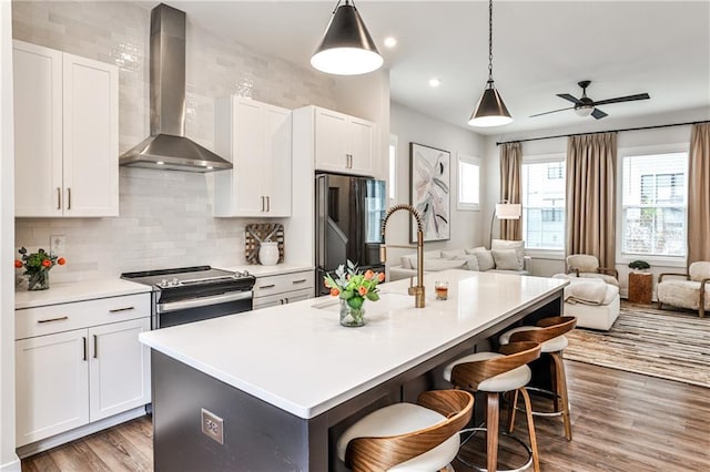 kitchen featuring stainless steel electric range oven, refrigerator with glass door, open floor plan, a kitchen island with sink, and wall chimney exhaust hood