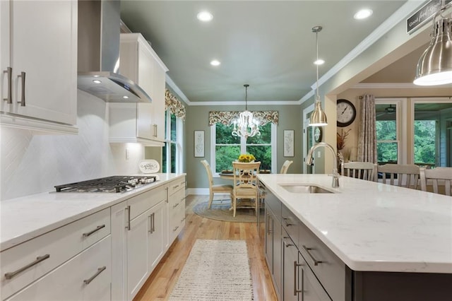 kitchen with wall chimney range hood, stainless steel gas stovetop, a sink, a center island with sink, and light wood finished floors