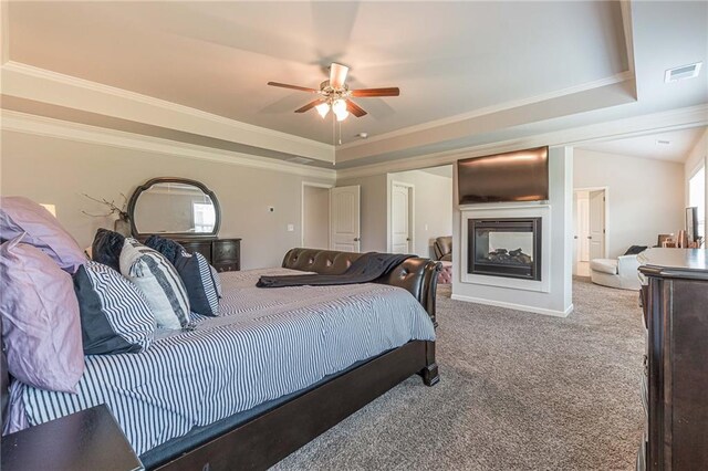 carpeted bedroom featuring ceiling fan, crown molding, a tray ceiling, a multi sided fireplace, and multiple windows