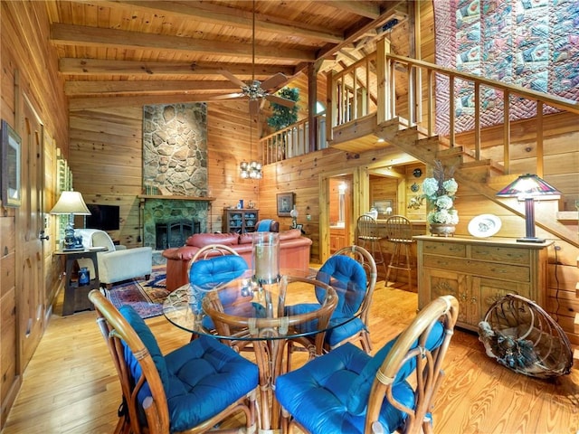 dining room featuring wood walls, light hardwood / wood-style floors, vaulted ceiling with beams, ceiling fan, and a stone fireplace