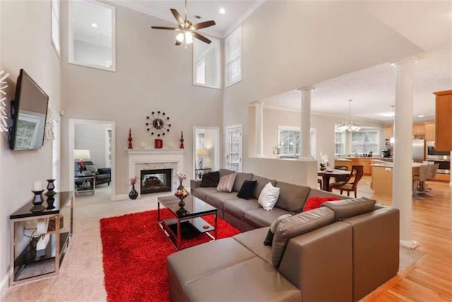 living room featuring a high end fireplace, ceiling fan with notable chandelier, light wood-type flooring, a towering ceiling, and decorative columns