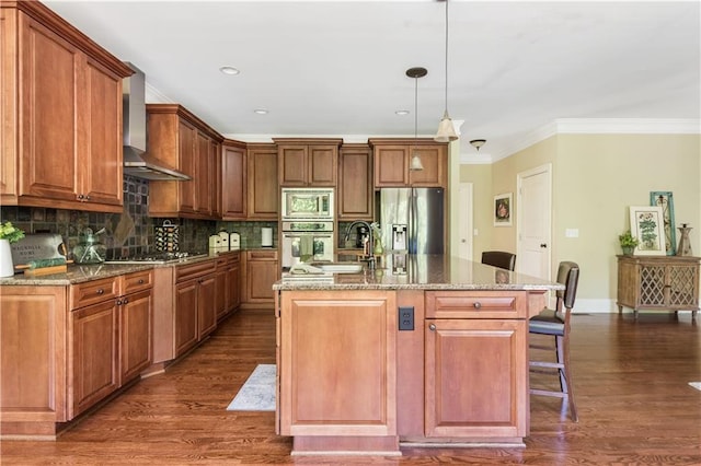 kitchen featuring dark wood-type flooring, wall chimney range hood, an island with sink, decorative light fixtures, and appliances with stainless steel finishes