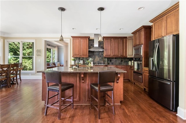 kitchen featuring wall chimney exhaust hood, stainless steel appliances, dark hardwood / wood-style floors, decorative light fixtures, and a center island with sink