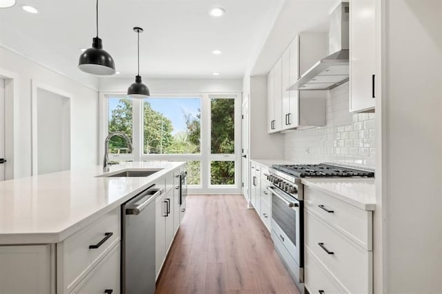 kitchen with light wood-type flooring, stainless steel appliances, hanging light fixtures, wall chimney exhaust hood, and white cabinets