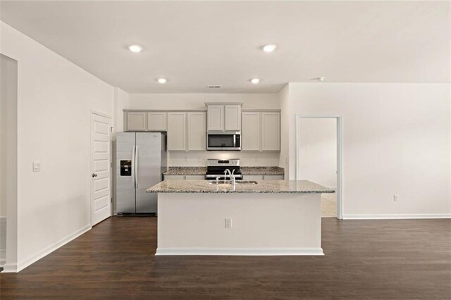 kitchen with dark wood-type flooring, sink, light stone counters, a center island with sink, and appliances with stainless steel finishes