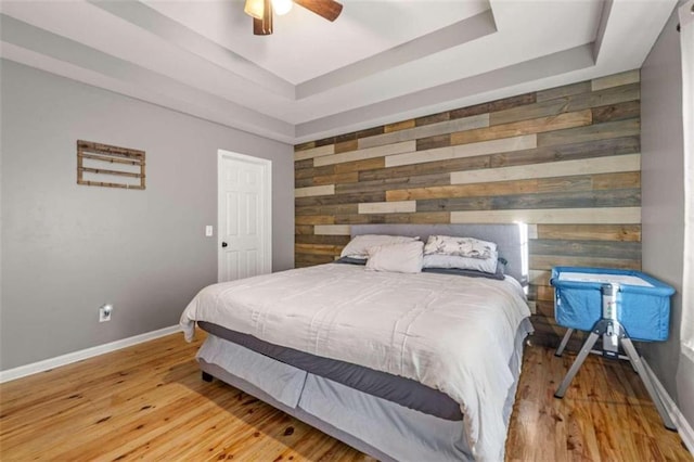 bedroom with wooden walls, ceiling fan, a tray ceiling, and light wood-type flooring